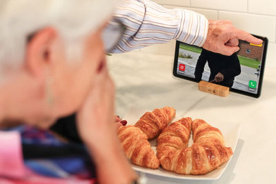 An elderly woman in a kitchen uses a smartphone on a wooden stand to video call her child, while croissants sit on a plate in the foreground.