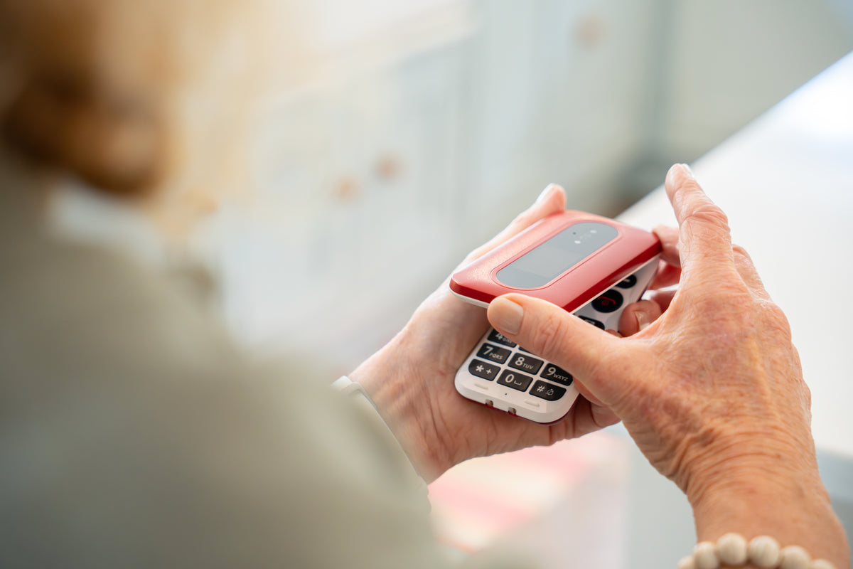 A close-up of a person's hands holding a small red and white flip phone with a keypad, shown in a softly blurred indoor setting.